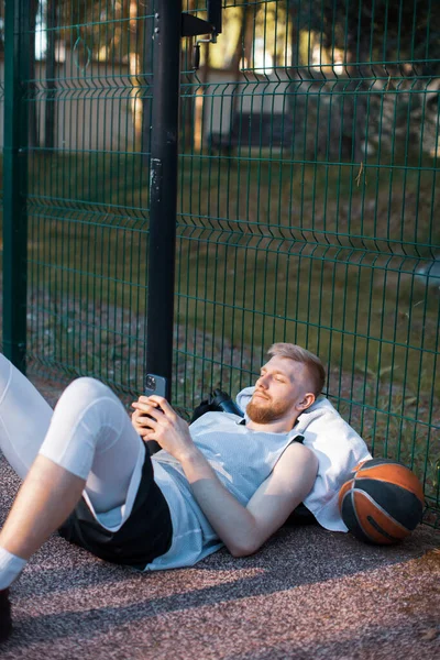 Jugador de baloncesto atlético macho acostado relajándose con el teléfono después de entrenar al aire libre en la cancha deportiva — Foto de Stock