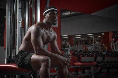 Afrikalı Amerikalı sporcu kaslı adam spor bankında bir şişe suyla oturuyor, antrenman sırasında kırılıyor.