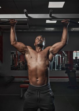 Çıplak gövdeli atletik Afrikalı Amerikalı adam barfiks çekiyor ya da spor salonunda egzersiz yapıyor.