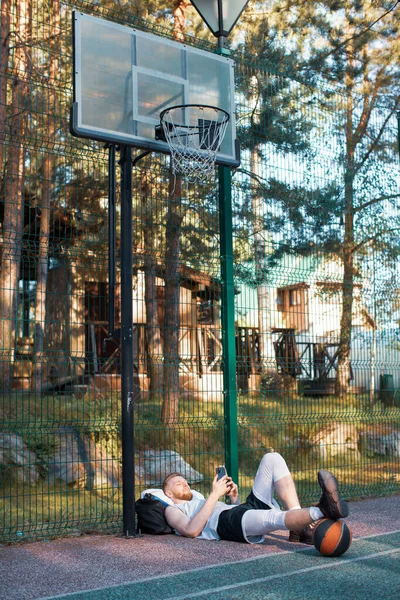 Jogador de basquete masculino usando telefone aplicativo esportivo relaxante após o treino ao ar livre sob arco de basquete — Fotografia de Stock