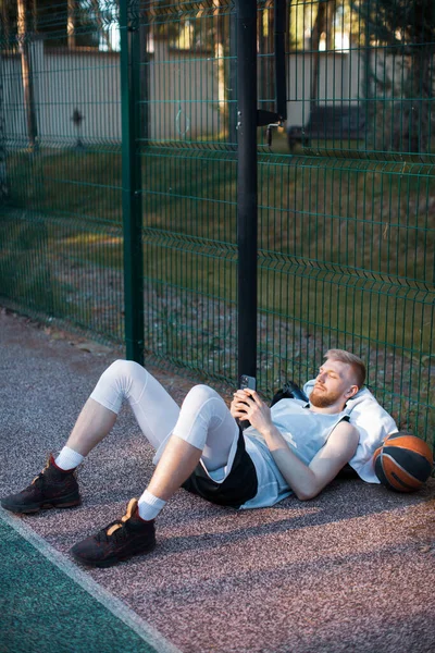 Баскетболист спортивный мужчина лежит расслабляясь с телефоном, смотрит новости в социальных сетях после тренировки на спортивной площадке — стоковое фото