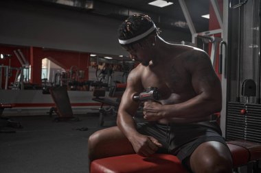 Atletik Afrikalı Amerikalı bir adam elle masaj yapıyor. El masajı tabancası, spor salonunda egzersiz sonrası iyileşme rutinleri.