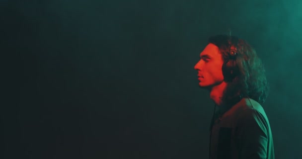 Танцюючий стильний молодий хлопець в навушниках слухати музику, насолоджуючись червоним студійним світлом з димом, копіювати простір . — стокове відео