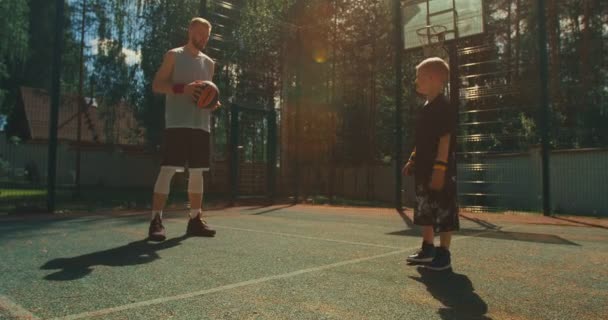Koszykarze. chłopiec pokaż na rękach stojak ćwiczenia z sportowe męski instruktor na boisku do koszykówki w słońcu — Wideo stockowe