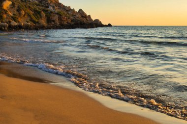 Mavi su turuncu gökyüzü, kum ve kayalar ile çevrili Golden Bay, Malta gün batımında.