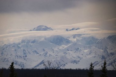 Alaska 'daki Denali Otoyolu' nda bir bahar günü karlı bir dağın tepesini kaplayan açık bulutlar..