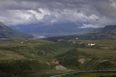 Alaska Trans-Alaska Boru Hattı yeşil tepelerden geçiyor. Arkasında dağlar var. Alaska 'nın iç kesimlerinde bulutlu bir yaz gününde..