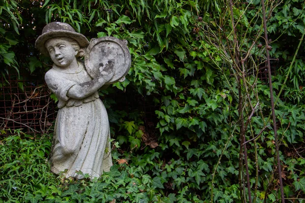 Old white weathered statue of a girl playing a tambourine surrounded with ivy and copy space