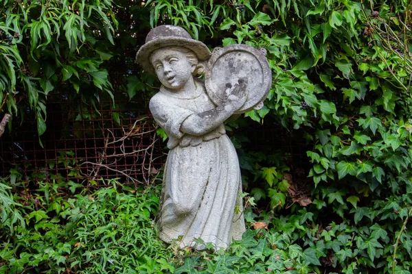 Old white weathered statue of a girl playing a tambourine surrounded with ivy