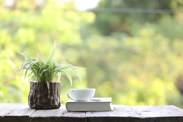 notebook and white coffee cup with plant in small wooden pot on wooden table at outside