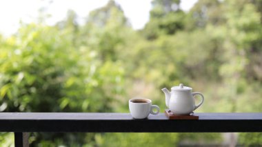 Balkon çitinin üstünde doğa manzaralı küçük çay fincanı ve çaydanlık.