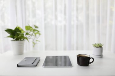 Not defterli tablet, kalem ve kahve fincanı yeşil bitkilerle beyaz masada.