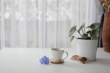 Kahve fincanı bitkisel ve kahverengi güneş gözlüklü beyaz masasında mor çiçekli ve perdenin arkasını gören.