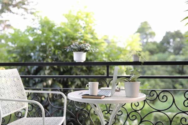 Coffee cup mug with laptop and notebook on white metal table decorated with plant pot outdoor balcony view 