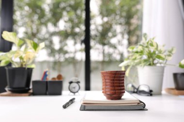 Kahverengi ahşap çay fincanı, defterler ve beyaz ahşap masa dekorunda bitkiler ve saatle birlikte kalem. 