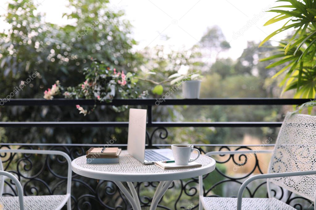 Coffee cup with laptop and notebook on white metal table decorated with plant pot outdoor balcony view