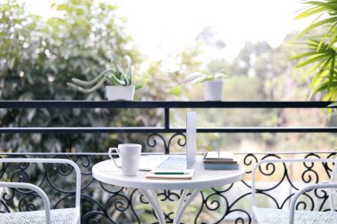 dizüstü bilgisayar ve kahve fincanı üzerinde beyaz metal bir masa üzerinde bitki otuyla süslenmiş balkon manzaralı.