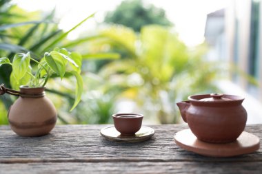 Kahverengi toprak çay takımı yemyeşil arka planlı, ahşap bir masaya yerleştirilmiş.