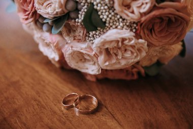 Altın alyanslar ahşap bir masanın üzerinde, gelinin gül buketinin arka planında yatıyor. Düğün konsepti