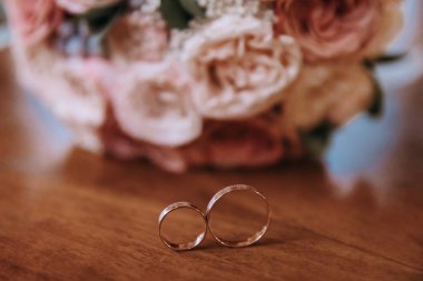 Altın alyanslar ahşap bir masanın üzerinde, gelinin gül buketinin arka planında yatıyor. Düğün konsepti