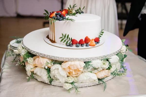 A beautiful wedding cake, decorated with fruits, raspberries, strawberries and blueberries. The bottom of the cake is decorated with rose flowers. Confectionery on the table. Holiday concept