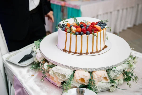 A beautiful wedding cake, decorated with fruits, raspberries, strawberries and blueberries. The bottom of the cake is decorated with rose flowers. Confectionery on the table. Holiday concept