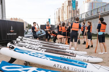 A group of young girls and boys stand in light wetsuits and equipment for sup surfing on land, the instructor gives instructions to beginners. A surf station. Russia, Saint Petersburg, 6.06.2021