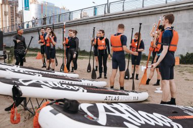 A group of young girls and boys stand in light wetsuits and equipment for sup surfing on land, the instructor gives instructions to beginners. A surf station. Russia, Saint Petersburg, 6.06.2021