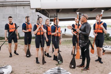 A group of young girls and boys stand in light wetsuits and equipment for sup surfing on land, the instructor gives instructions to beginners. A surf station. Russia, Saint Petersburg, 6.06.2021