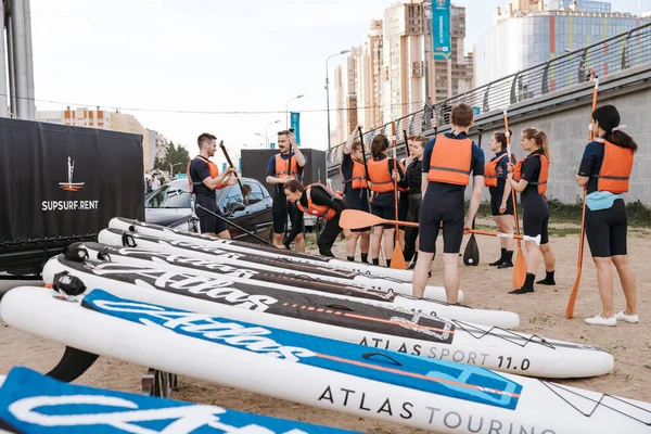 A group of young girls and boys stand in light wetsuits and equipment for sup surfing on land, the instructor gives instructions to beginners. A surf station. Russia, Saint Petersburg, 6.06.2021