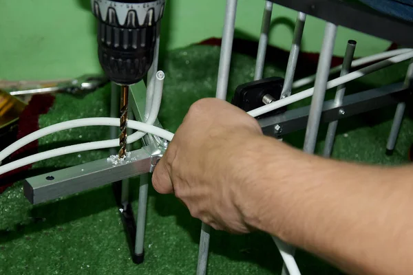 On a green background, surrounded by various spare parts, a man drills with a drill in an aluminum strip, antenna. The man drills holes for the antenna.