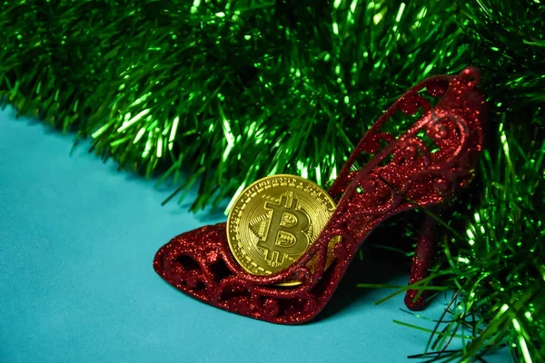 Die Bitcoin-Münze steckt in einem roten Frauenschuh. Weihnachtliches Lametta und ein roter Frauenschuh mit einer Goldmünze aus Kryptowährung. Stockbild