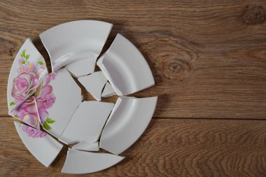 Shards of a broken plate on the wooden floor. Broken white ceramic plate on the wooden floor. Broken dishes. Top view of a damaged ceramic plate. Empty space for text. clipart