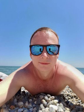 Güneş gözlüklü ve kulağında küpe olan bir adam deniz kıyısındaki çakıl taşı plajında güneşleniyor.