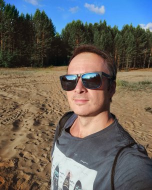 Ormandaki kumlu bir arazide tişörtlü ve güneş gözlüklü bir adamın selfie portresi.