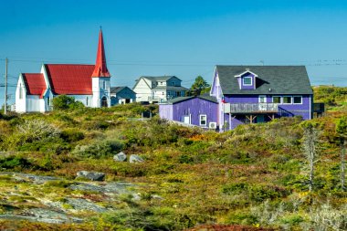 Peggy 's, Cove, NS - CA - 29 Sep 2025 Canlı mor bir ev ve kırmızı çatılı beyaz bir kilise açık mavi gökyüzünün altında kayalık, yeşil bir yamaçta kurulmuştur..