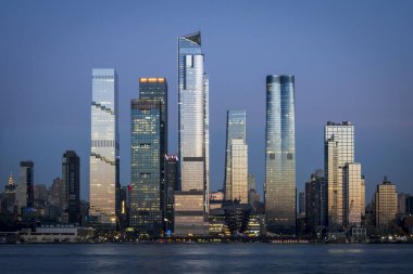 Manhattan Batı Yakası gökdelenleri alacakaranlıkta Hudson Nehri boyunca yükseliyor. Parlak cam kuleler, canlı şehir ışıkları ve New York 'un dinamik kentsel ufkunu şekillendiren modern deniz kenarı gelişmeleri ile.