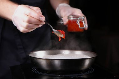 Kremalı havyar sosu hazırlama. Koruyucu eldiven giyen bir aşçı tavaya kırmızı havyar ekler. Siyah arkaplandaki fotoğraf. Tanınmayan kişi.