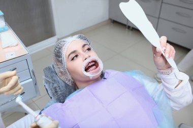 Dişçi randevusundaki genç bir kız aynada kendine bakıyor. Profesyonel ağız temizliği ve diş temizliği. Dişeti ve sakız hastalıklarının önlenmesi. Ağızda retraktör var..
