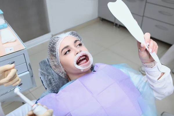 Een jong meisje bij een tandartsafspraak kijkt naar zichzelf in de spiegel. Professionele mondhygiëne en tandreiniging. Preventie van cariës en tandvleesziekten. Retractor in de mond. — Stockfoto