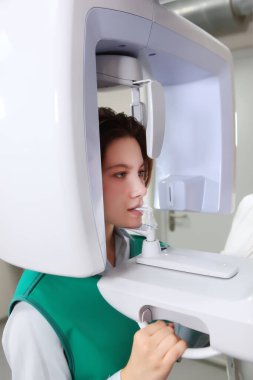 Röntgen odasında yeşil koruyucu yelek giyen bir hasta var. Diş kliniğinde röntgen cihazları. Modern tıp. Dişlerinin panoramik röntgeni. Yan görünüm.