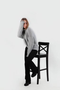 Bir kız, donmuş ellerini yün bir kazakla ısıtıyor. Siyah pantolonlu bir kız siyah bir sandalyeye oturur. Soğuk mevsim için doğal yün giysiler. Hafif arkaplan. Uzayı kopyala Dikey fotoğraf.