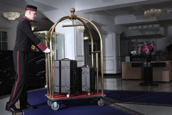 A uniformed doorman pushes a luggage cart .Several leather suitcases stand on a luggage cart. Hotel service.The holiday season. Free space.