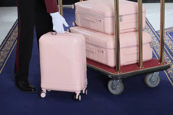 A uniformed doorman moves a cart with pink suitcases. A few suitcases on a luggage cart at the hotel. Doorman\'s job.Concept of hotel service.Luggage trolley at the hotel.