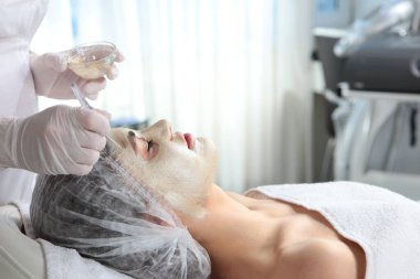 Bir kozmetikçi, bir kadının yüzüne altın kozmetik maske takmak için fırça kullanır. Güzellik ve gençliğin korunması kavramı. Bir kozmetoloji kliniğinde prosedür. Yan görünüm.