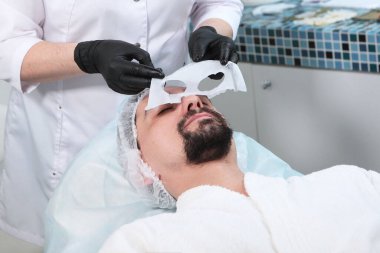 Bir kozmetikçi sakallı genç bir adam için maske yapar. Erkekler kozmetik kliniğinde bir adam. Modern bir adam..