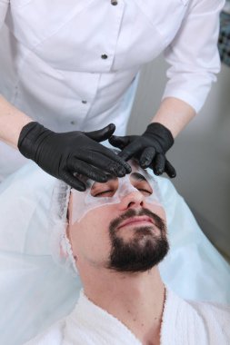 Bir kozmetikçi sakallı genç bir adam için maske yapar. Erkekler kozmetolojisi kliniğinde bir adam. Gençliği koruma ve yüz derisi ile ilgilenme kavramı.