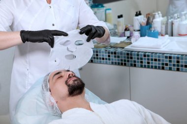 Güzellik uzmanı nemlendirici yüz maskesi tutuyor. Adam kozmetoloji kliniğinde yüz tedavisi yapıyor. Erkek kozmetolojisi. Gençliğin korunması ve cilt bakımı kavramı.