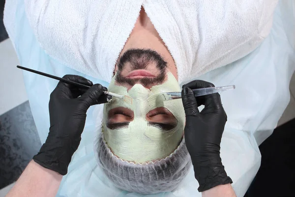 The beautician applies a nourishing mask to the man's face. A man doing facial procedures in a cosmetology clinic. Men's cosmetology. The concept of preservation of youth and skin care of the face.Top view.