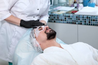 Kozmetik uzmanı alnında ve sakallı bir adamın gözlerinin etrafında maske yapıyor. Erkekler kozmetik bilimi kliniğinde. Gençliğin korunması ve yüz bakımı. Güzellik salonu kavramı..
